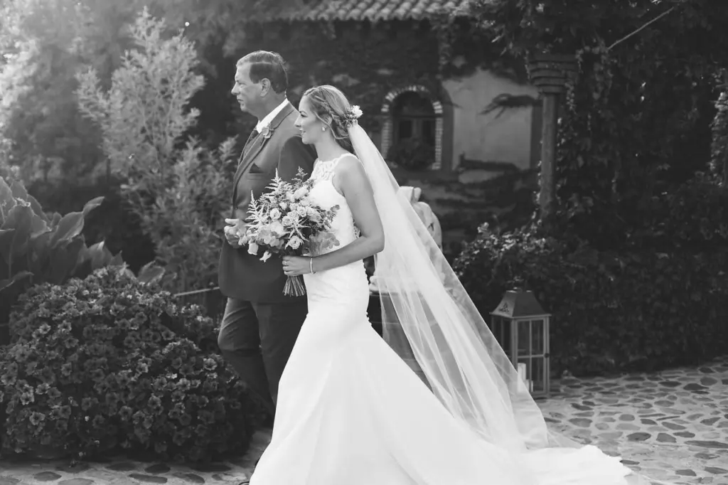 Novia con ramo y velo del brazo del padrino, en blanco y negro